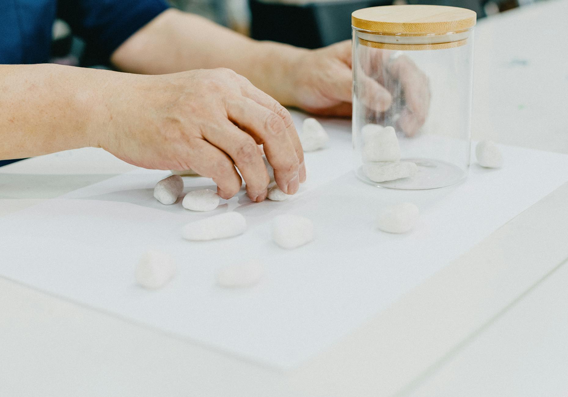 Dłonie układają białe kamienie i szklany słoik na kartce do rysunku